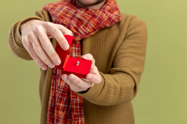 男人身穿休闲服 脖子上围着围巾的年轻人展示了红色盒子和订婚戒指 绿色背景下是情人节的概念订婚戒指天