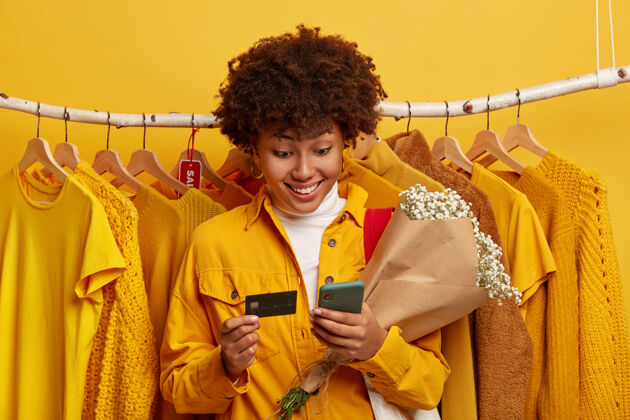 欢呼卷曲的黑皮肤女人使用信用卡和现代手机商店信贷微笑