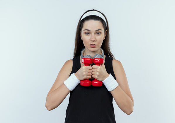 站立戴着头巾的年轻健身女 正脸举着哑铃站在白墙上锻炼健身年轻女人