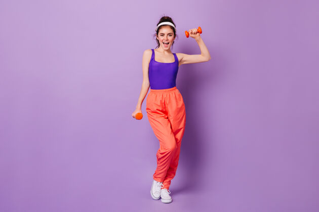 成人可爱的运动型女人 穿着80年代风格的亮丽健身服 微笑着用哑铃演示运动连体衣肖像移动