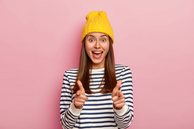 位置快乐的女人直指镜头 用手指枪的手势表达自己的选择 戴着黄色帽子和条纹休闲套头衫 条纹显示毛衣