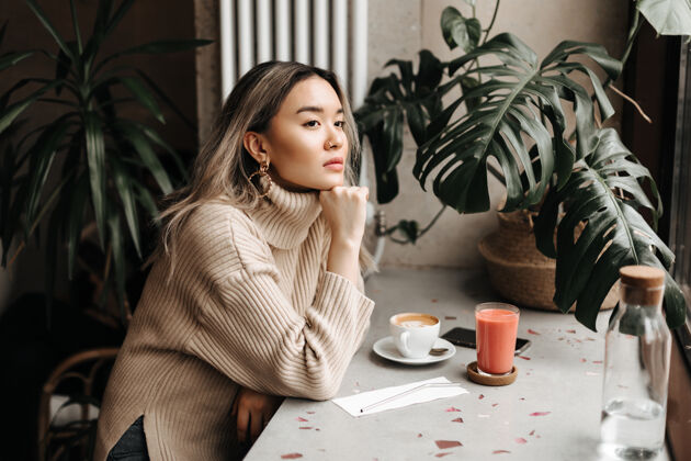 肤色穿着时尚米色毛衣的女人若有所思地看着远方 倚在桌上 端着一杯咖啡和新鲜果汁放松女士亚洲人