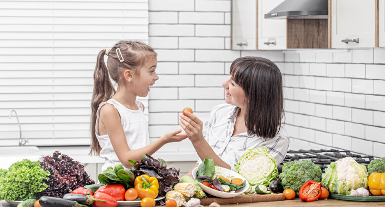 新鲜开朗的妈妈和女儿在一个明亮的现代厨房里用许多蔬菜做蔬菜沙拉自然烹饪配料