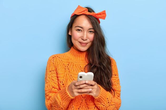 快乐快乐美丽的亚洲长发女人肖像 戴着头带和橙色毛衣 手持手机聊天 下载新应用程序科技女性日语