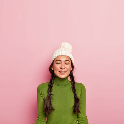 冬天高兴的黑发女孩闭上眼睛 想着和朋友愉快的见面 戴着白帽子和绿色高领毛衣 靠着粉色的墙站着胭脂衣服人