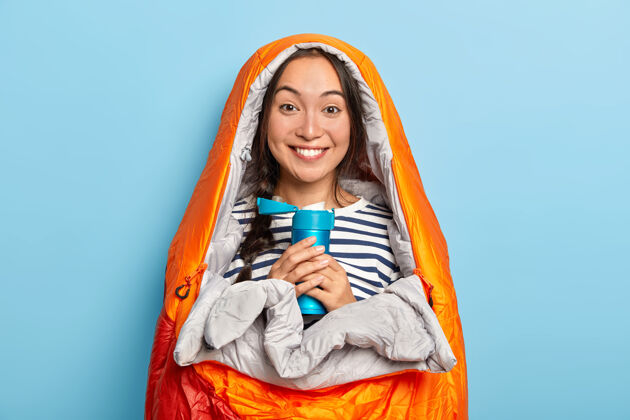 旅行年轻快乐的亚洲女人在睡袋里取暖 拿着装有热芳香饮料的蓝色烧瓶 感觉很放松 把业余时间花在大自然上 隔着蓝色的墙与世隔绝热水瓶茶旅行