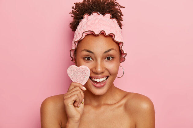 健康美丽的女人拥有健康的皮肤 用化妆棉擦脸 积极微笑 露出洁白的牙齿 戴着淋浴头带 下班后洗澡 隔离在粉红色的墙上裸体发型头带