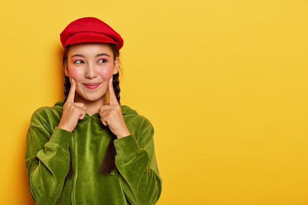 化妆年轻漂亮的亚洲模特把两个前指放在脸颊上 除了梦幻般的表情 妆容简约 头戴红色贝雷帽 头戴绿色天鹅绒连帽衫版面立场欢呼