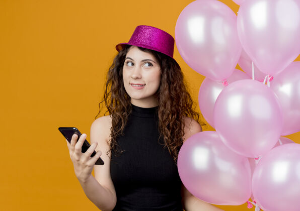 抱着一个年轻漂亮的女人 卷发 戴着节日礼帽 手里拿着一堆气球和智能手机 抬头思考着咬着嘴唇的生日派对概念站在橙色的墙上女人年轻束