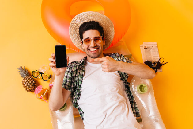 行李那个家伙躺在充气床垫上 上面放着菠萝 报纸和复古相机戴着帽子和眼镜的男人指着智能手机水果短裤包