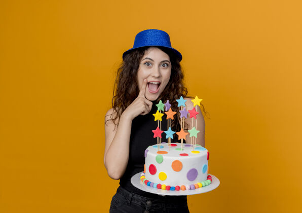 年轻一位年轻漂亮的卷发女士 戴着节日礼帽 手里拿着生日蛋糕 欢快地站在橙色的墙上抱着帽子看着
