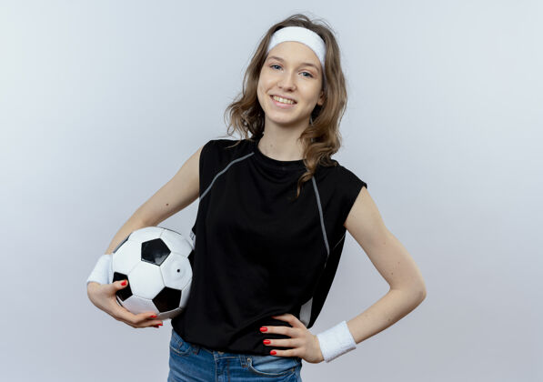 球身穿黑色运动服 头箍 手持足球 自信地站在白墙上的年轻健身女孩健身微笑女孩