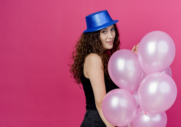 女人一个年轻漂亮的女人 一头卷发 戴着节日礼帽 手里拿着一堆气球 微笑着站在粉色的墙上站着气球空气