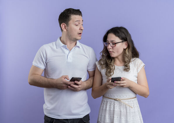 男人一对年轻夫妇手持智能手机站在蓝色的墙上互相监视女人站着移动