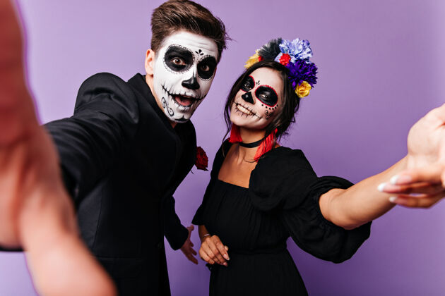 墨西哥有趣的年轻人和他的女朋友在他们的脸上带着微笑自拍在紫色工作室的万圣节化妆的顽皮夫妇的肖像男人花幽灵