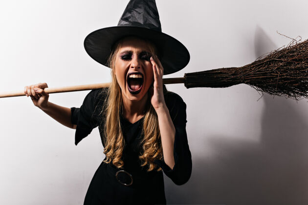 巫术一头金发的女巫在白墙上尖叫年轻的女巫师拿着她的魔法扫帚嘉年华女巫魔鬼