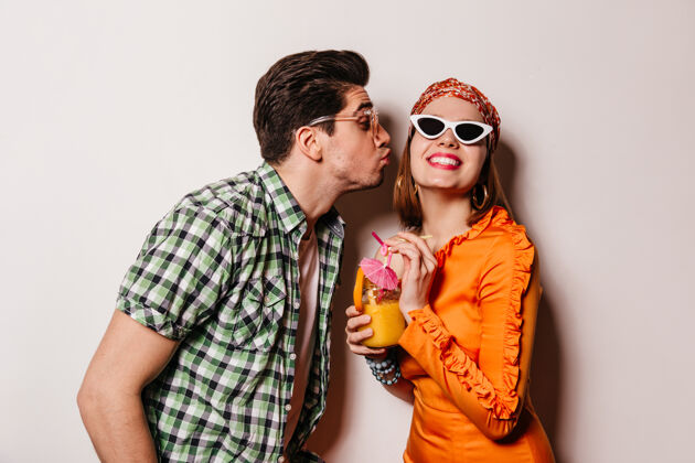 老式相机身穿格子衬衫的黑发男人亲吻他的女朋友戴着眼镜和橙色裙子的女士微笑着拿着鸡尾酒衬衫友谊漂亮