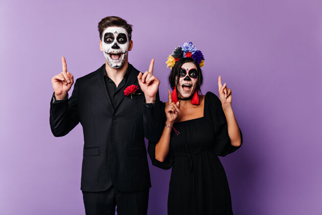 恐怖快乐的黑眼睛男孩和女孩情绪化地摆出姿势 伸出手指紫色墙上墨西哥风格的面部艺术的夫妇照片玫瑰文化化妆舞会