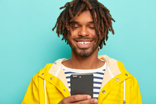一个一个戴着长发的笑脸男人的肖像 穿着黄色雨衣 用一部手机 隔离在蓝色背景上美国男士聊天
