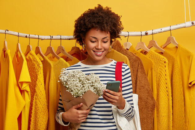 聊天漂亮快乐的非洲女人专注于智能手机 手里拿着漂亮的花束 开心地笑着 站在挂在衣架上的黄色衣服旁边顾客精品店设备
