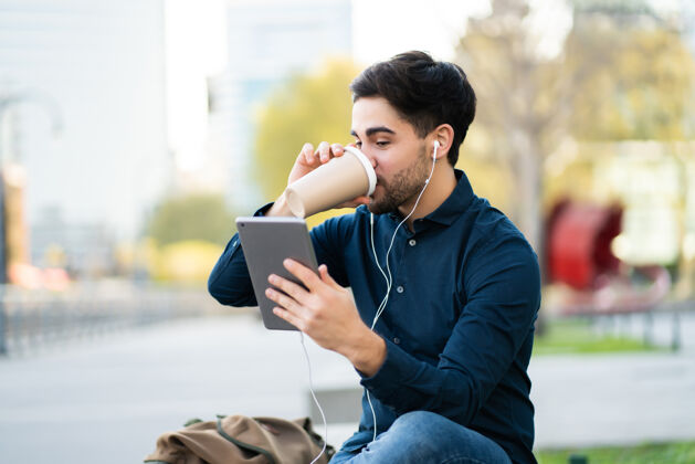 城市年轻人坐在户外的长椅上 通过数字平板电脑进行视频通话并喝咖啡的照片城市概念互联网触摸屏工作