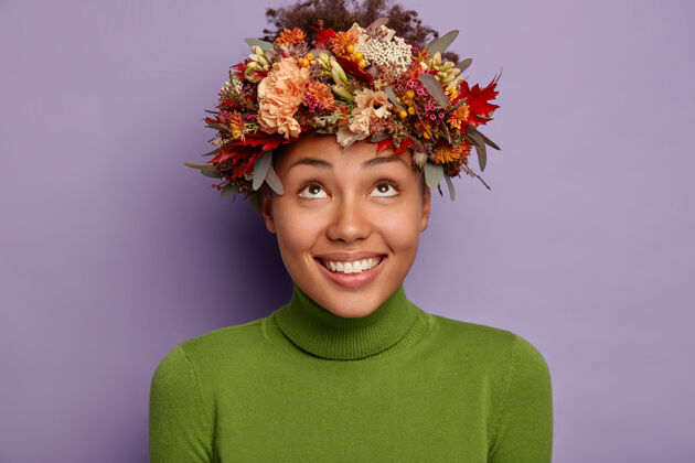 非洲美丽的秋日非洲女孩 带着喜悦的表情 看着用时令植物做成的美丽的秋日花环 穿着绿色高领毛衣 在紫色的背景下与世隔绝牙关向上休闲