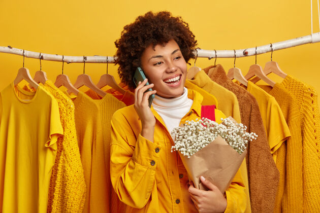 非洲穿着亮黄色夹克的快乐女人的照片 站在衣柜衣架上的衣服旁 准备出门 拿着花束通过手机给朋友打电话女性购物狂喜欢黄色消费主义欢呼时尚
