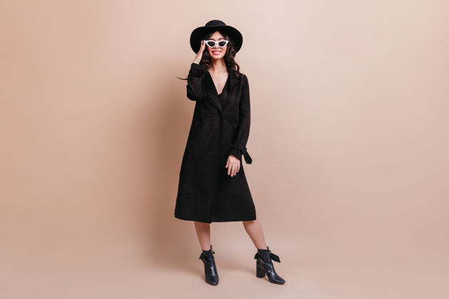 日本人戴着帽子和太阳镜的女人的全景图优雅的深色女人穿着外套站在米色背景上享受女士年轻