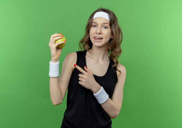 运动装身穿黑色运动服 头戴绿帽子的年轻健身女孩站在绿色的墙上 用手指着绿色的苹果微笑着手指微笑苹果
