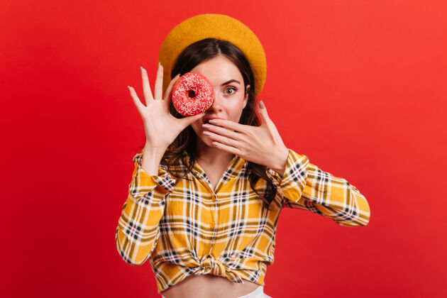 成人特写镜头中 一个惊讶的绿眼睛女孩拿着美味的甜甜圈戴贝雷帽和衬衫的女士捂着嘴年轻法国时尚
