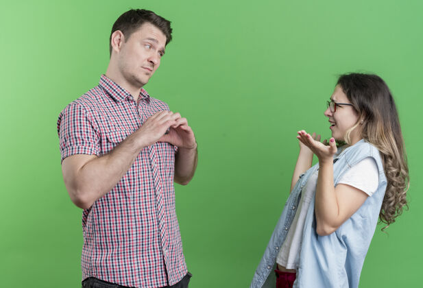 心一对年轻夫妇穿着休闲服的男人和女人伤心的男人向站在绿色墙壁上的她不高兴的女友展示心形情侣显示姿态