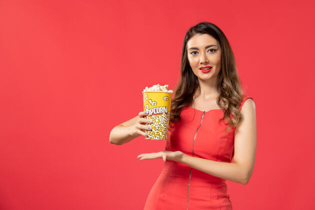 电影正面图身穿红色衬衫的年轻女性手持爆米花包 在红色表面微笑漂亮女士包装