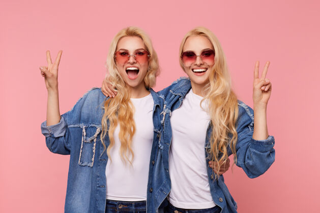 双胞胎快乐的年轻迷人的长发金发女人戴着太阳镜举手致胜的手势 愉快地看着镜头迷人的微笑 孤立在粉红色的背景下T恤休闲红色