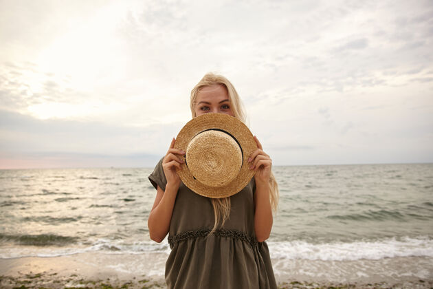 海岸积极的年轻蓝眼睛迷人的白头女性保持她的船帽在自己面前 同时高兴地看着摄像机 在海滩背景隔离提高手姿势