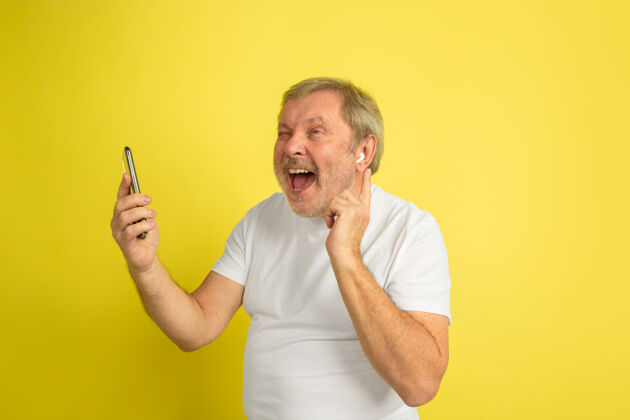 养老金戴着耳机和智能手机唱歌黄色工作室背景上的白种人肖像穿着白衬衫的漂亮男模时尚手机半