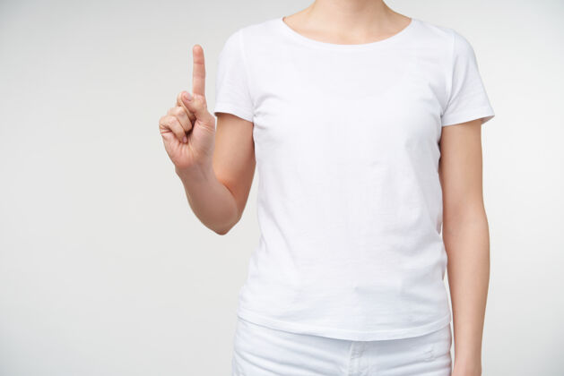 身体摄影棚照片中的年轻女子身穿白色t恤 在白色背景上摆姿势 一边数一边举起食指手和手势的概念手臂计数语言