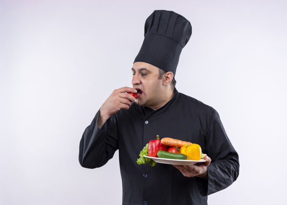 站着男厨师身穿黑色制服 头戴厨师帽 手拿新鲜蔬菜夹西红柿的盘子站在白色背景上白色男制服