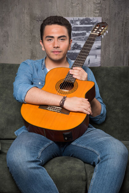 玩年轻的吉他手拿着一把漂亮的吉他坐在沙发上高品质的照片和弦男孩成人