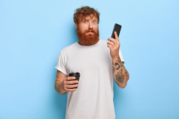 外卖困惑的大胡子红发男人拿着手机 接到陌生人的电话 通过手机听到可怕的尖叫声 喝外卖咖啡 穿着白色休闲t恤工作姜压力