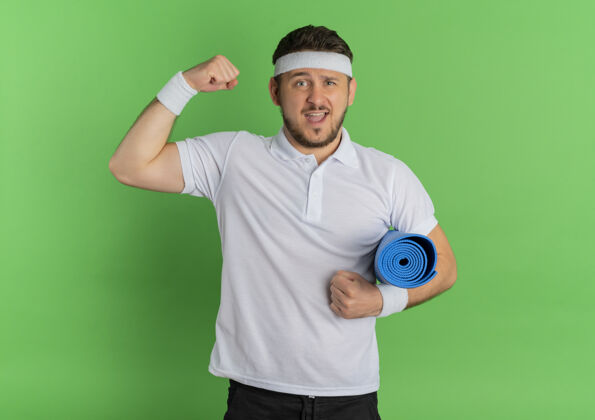 垫子身穿白衬衫 头箍 手持瑜伽垫 举起拳头 展示肱二头肌的年轻健身男子 站在绿色背景上 展现快乐和积极的赢家理念举行绿色显示