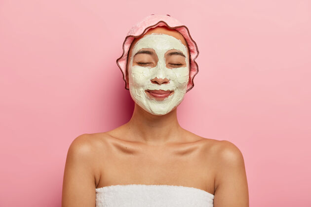 年轻高兴的亚洲女人戴着粘土净化有机面膜在脸上 有美容程序在水疗沙龙 戴着玫瑰色软保护淋浴帽按摩泥健康