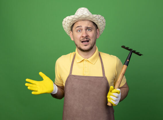 蔓延年轻的园丁穿着连体衣 戴着帽子 戴着工作手套 拿着小耙子看着前面 困惑地伸开手臂站在绿色的墙上迷你手臂人