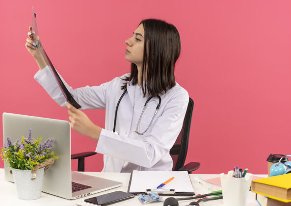 周围年轻的女医生穿着白大褂 脖子上戴着听诊器 正坐在桌旁看肺部x光片 一脸严肃 笔记本电脑盖在粉红色的墙上脸坐着严重