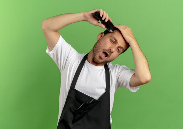 工作穿着围裙的理发师站在绿墙上用理发机剃着头发 看上去很自信男人站摆姿势