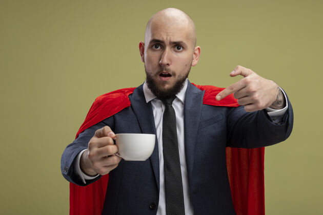 困惑超级英雄 留着胡子 披着红斗篷的商人 拿着一杯咖啡 用食指指着它 站在淡绿色的墙上 困惑不已胡子超级英雄站着