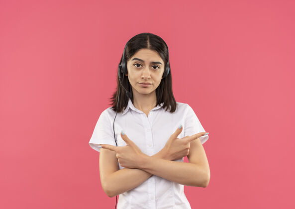 脸穿着白衬衫 戴着耳机的年轻女孩 面带严肃的表情 手指着站在粉色墙上的两侧侧身衬衫站