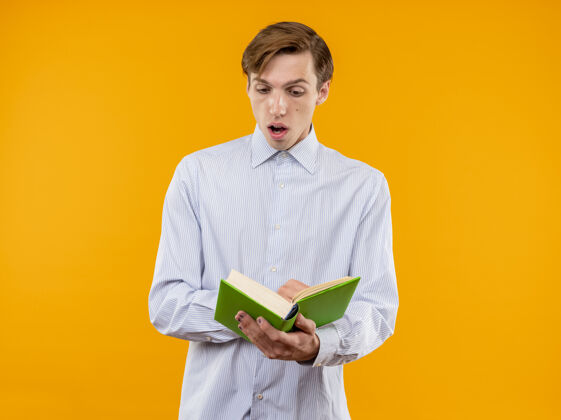 开放一个穿着白衬衫的年轻人站在橘色的墙上 手里拿着一本打开的书 感到惊讶和惊奇惊奇阅读年轻
