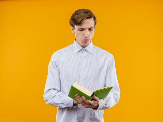 男人身穿白衬衫的年轻人拿着打开的书 严肃地站在橙色的墙上看着书抱着橙色脸