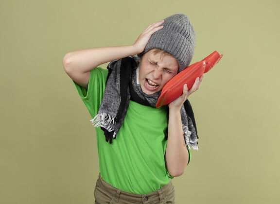 光生病的小男孩穿着绿色t恤 戴着暖和的围巾和帽子 感觉不舒服 拿着水瓶取暖 站在轻墙上头痛得厉害温暖小疾病