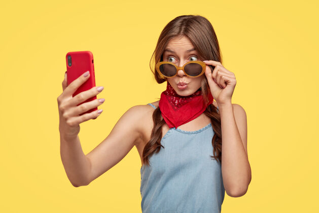 青少年独立拍摄的时尚少女撅着嘴唇 戴着时髦的太阳镜 脖子附近戴着红色的头巾 拿着智能手机 自拍肖像 享受闲暇时光 站在黄色的墙上白种人拍照黑发